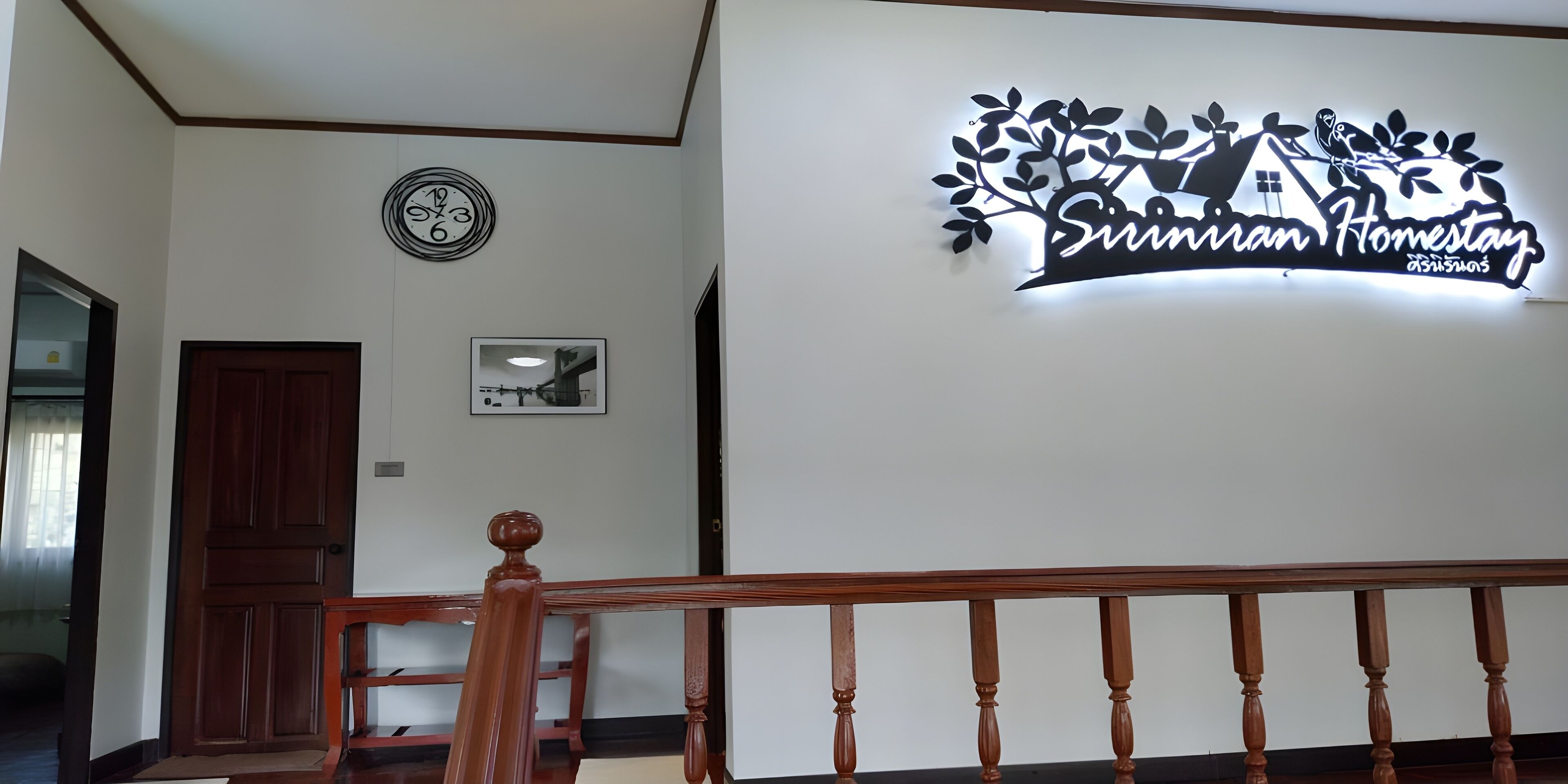 A hotel interior with a wooden staircase, a clock, a framed picture, and a sign reading "Sirimiran Homestay".
