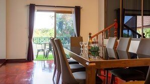 A dining area with a wooden table and chairs, a glass-top table, and a view of the outdoors through a large window. - Siriniran Homestay (Sattahip)