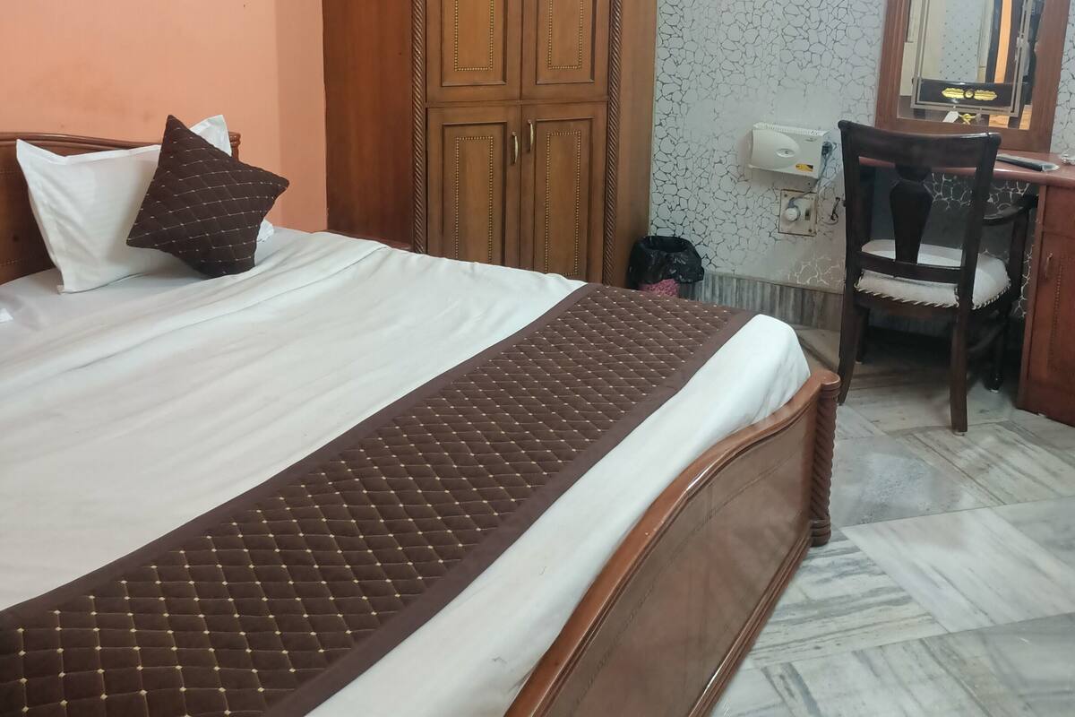 A hotel room with a bed, a wardrobe, a desk with a chair, an air conditioning unit, and a mirror.