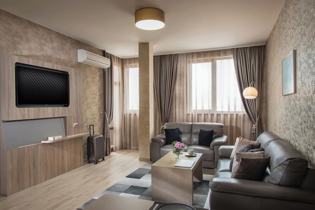 A modern hotel room with a leather sofa, a wooden coffee table, a flat-screen TV mounted on the wall, and a large window with curtains.