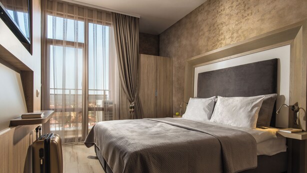 A hotel room with a large bed, a suitcase, a desk with a lamp, and a view of the city through large windows. - Sevtopolis Apartments (Kazanlak)