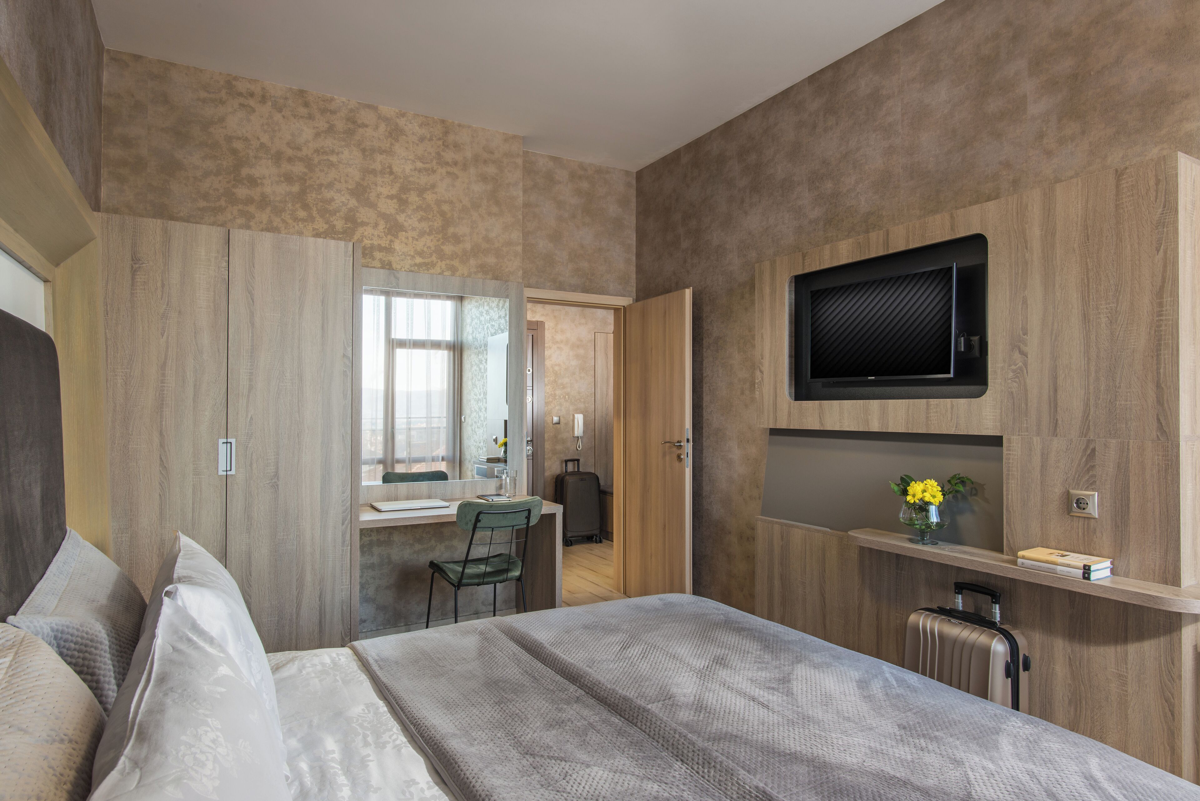 Een moderne hotelkamer met een bed, een bureau, een stoel, een aan de muur bevestigde televisie en een koffer op de vloer.