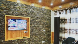 Una habitación con pared de piedra, puerta de madera, televisor montado en la pared y ventana con cortinas.