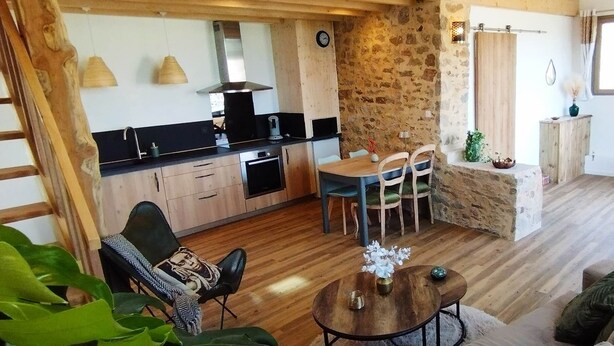A cozy living room with a wooden floor, a dining area with a table and chairs, a kitchen with a stove and cabinets, and a stone wall.