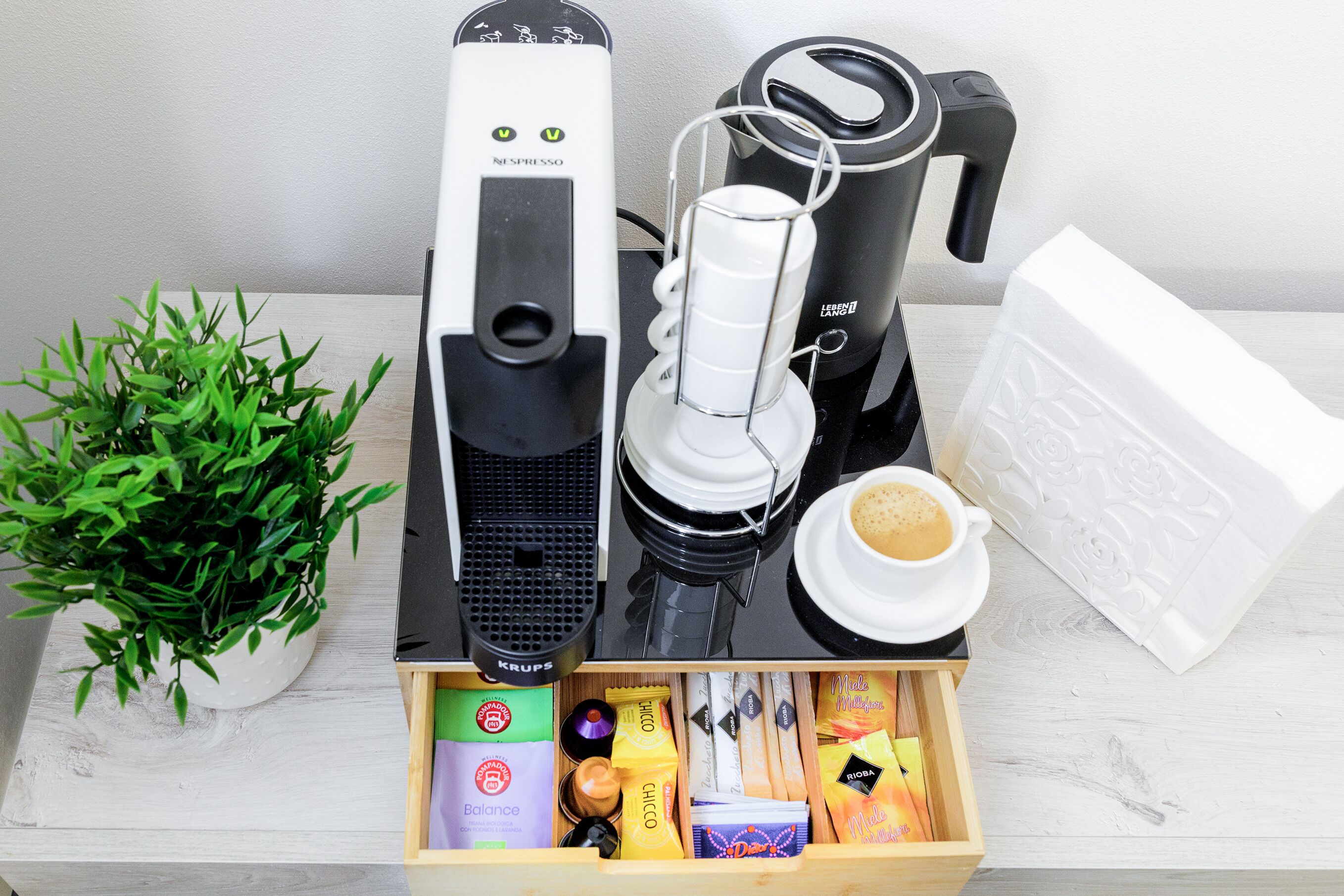 En kaffebryggare, en kaffekanna, en kopp kaffe, en växt och en förpackning med kaffekapslar.