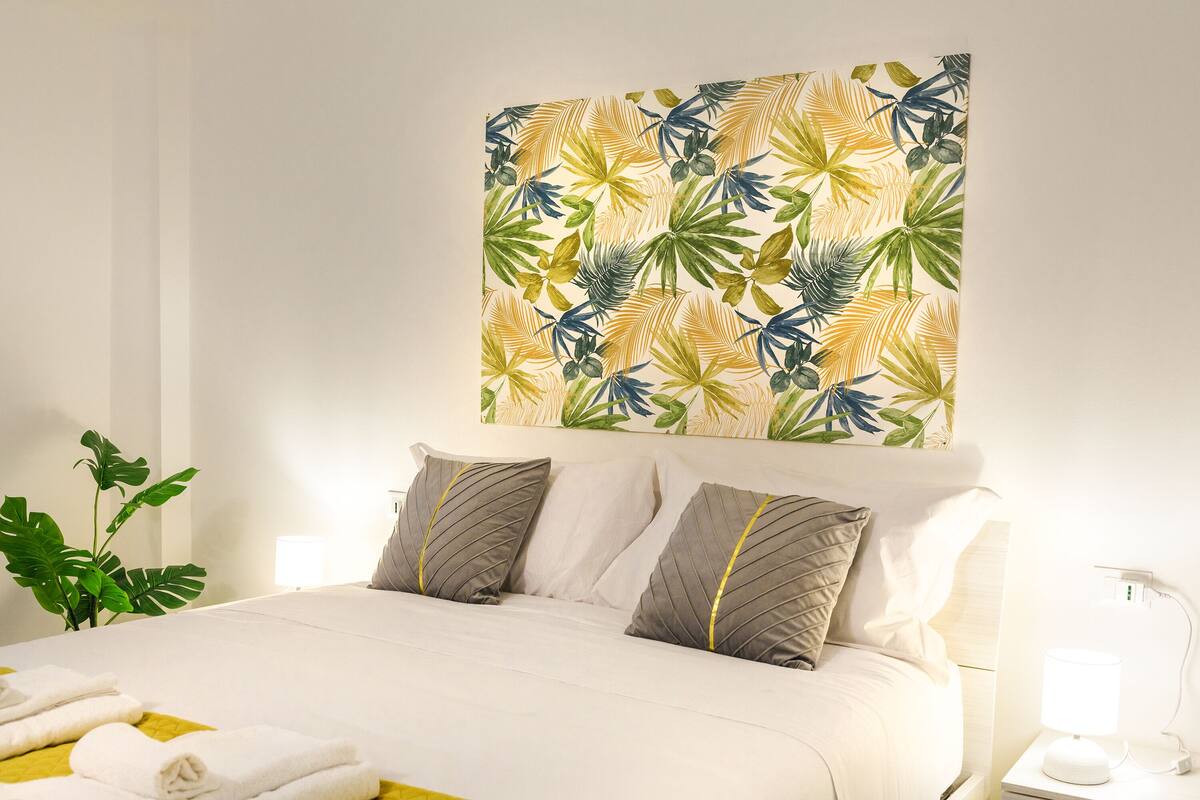 A hotel room with a bed, pillows, a bedside table with a lamp, and a wall with a tropical leaf pattern.