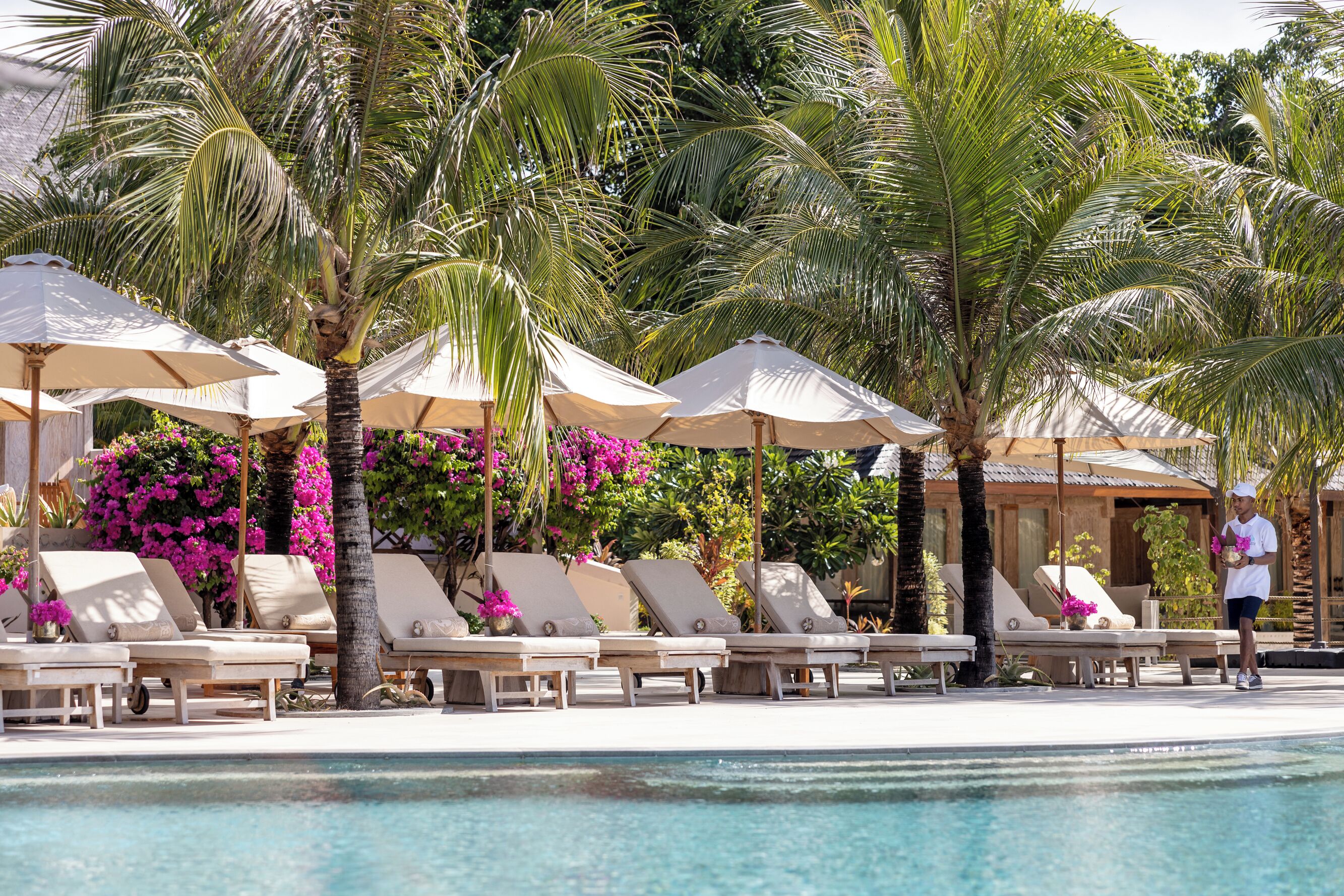 A poolside area with lounge chairs and umbrellas, surrounded by palm trees and flowering plants.