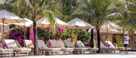A poolside area with lounge chairs and umbrellas, surrounded by palm trees and flowering plants.