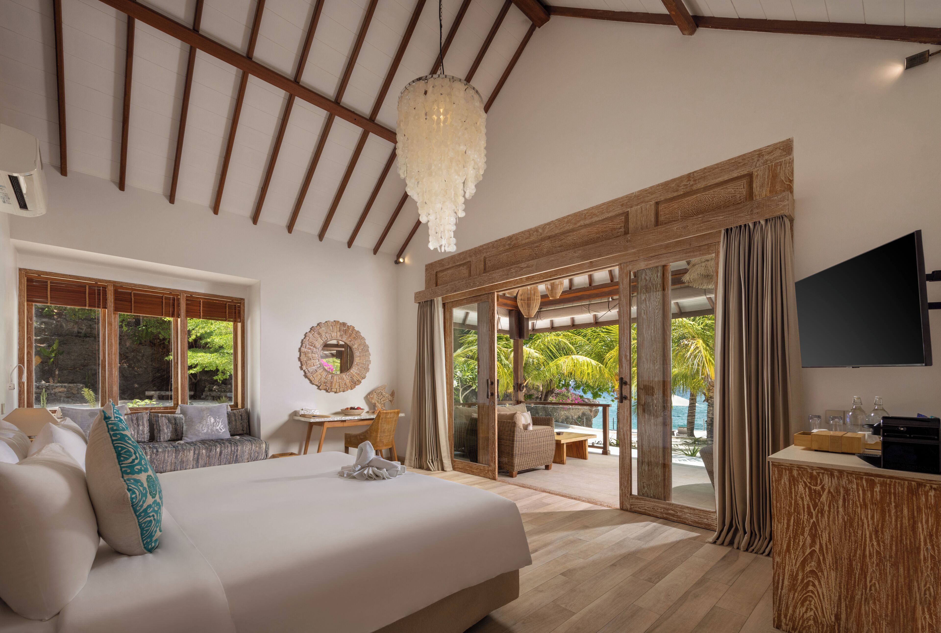 A bedroom with a large bed, a desk, a chair, a TV, and a view of the outdoors.
