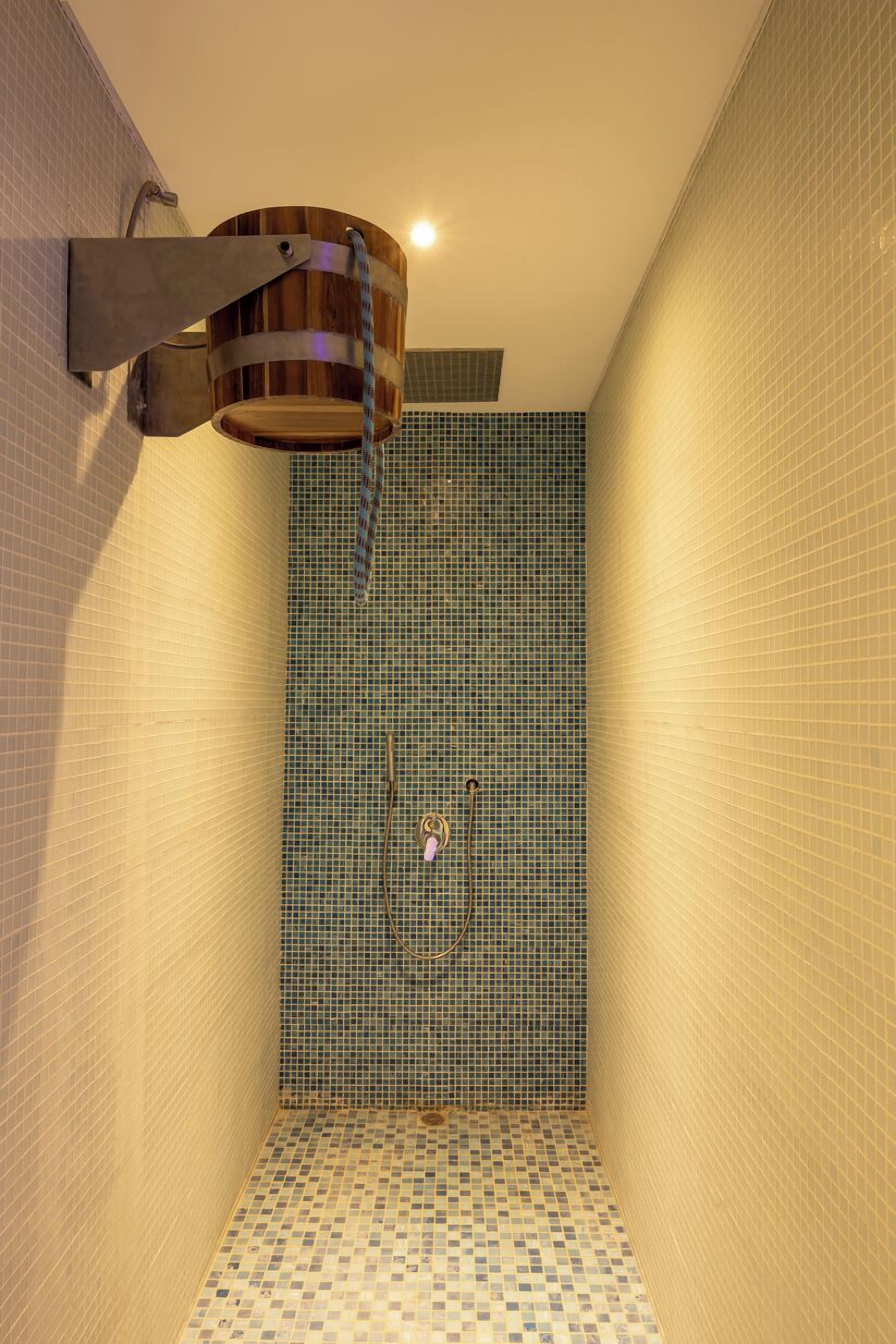 A narrow shower with a wooden bucket mounted on the wall, tiled walls, and a tiled floor.