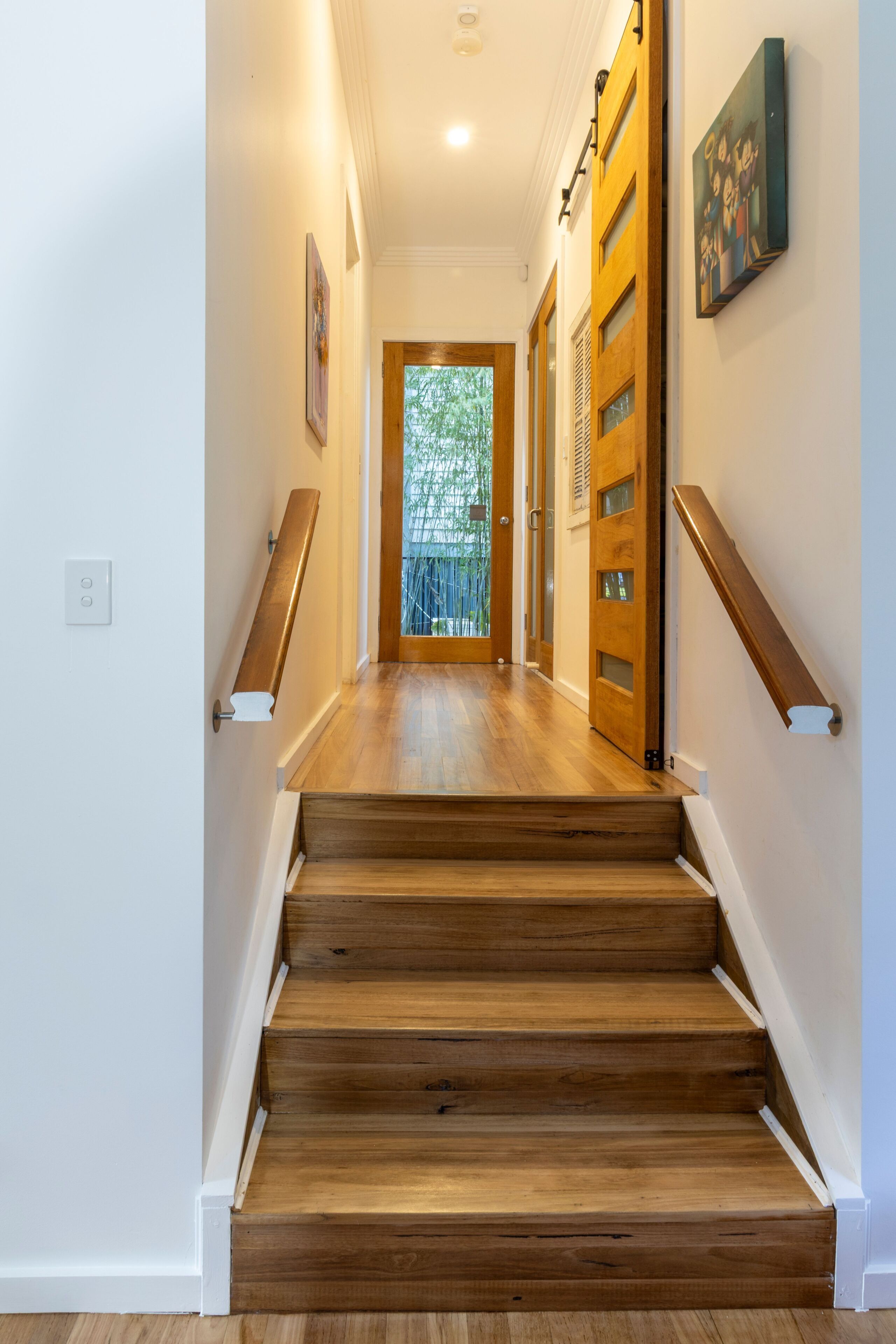 Un escalier en bois mène à une porte dotée d’un panneau de verre, le tout étant encadré par des murs blancs et un tableau.