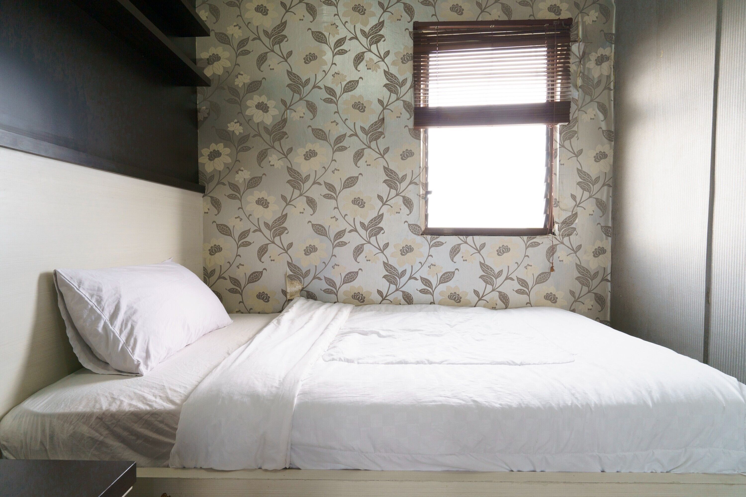 A single bed with white bedding in a room with floral wallpaper and a window with blinds.