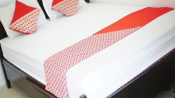 Een net opgemaakt bed met witte lakens en een deken met een rood-wit patroon. Er liggen twee rode kussens met een wit dessin.