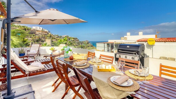 A rooftop terrace with a table set for dining, lounge chairs, and a grill.