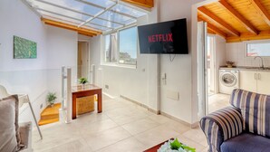 A modern living room with a large TV screen, a sofa, a dining area, and a kitchen with a washing machine.