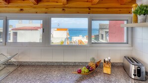 A kitchen with granite countertops, a toaster, a fruit bowl with grapes and a pineapple, and a view of the sea through large windows.