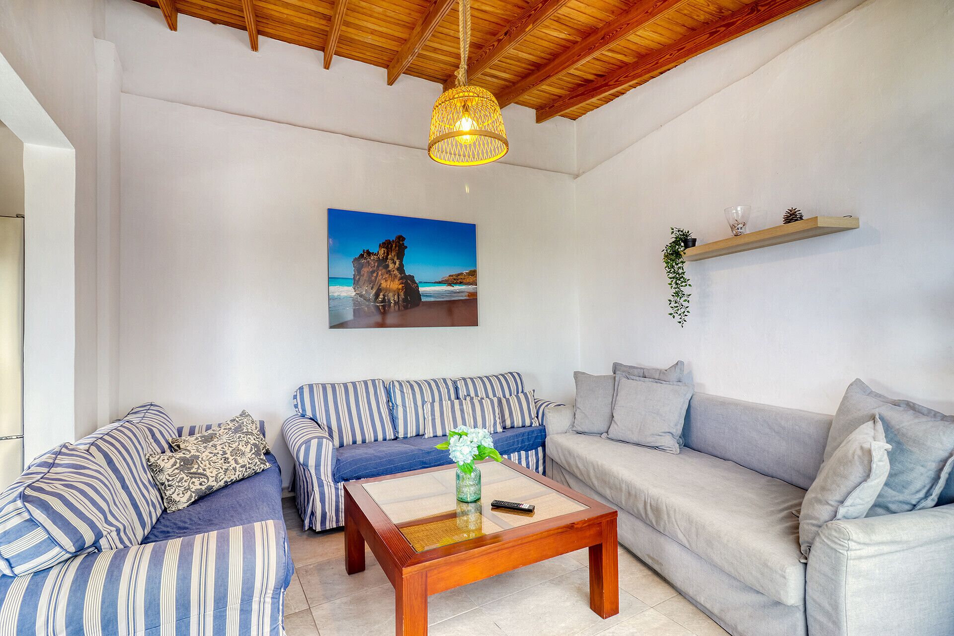 Una sala de estar con techo de madera, un cuadro de una escena costera, un conjunto de sofás, una mesa de centro y una planta en un estante.