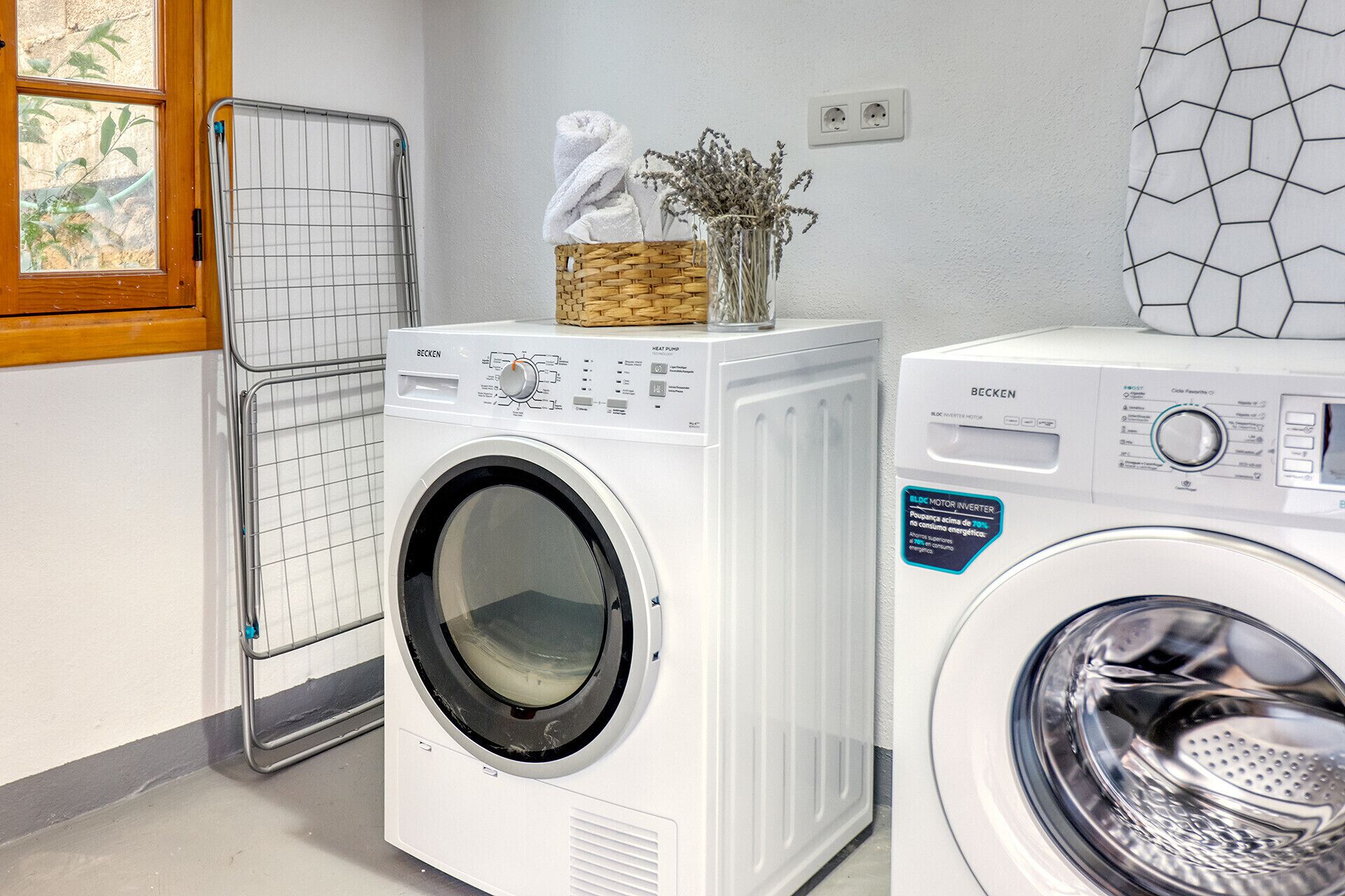 A laundry area with two white front-loading washing machines, a drying rack, and a window with a view of greenery.