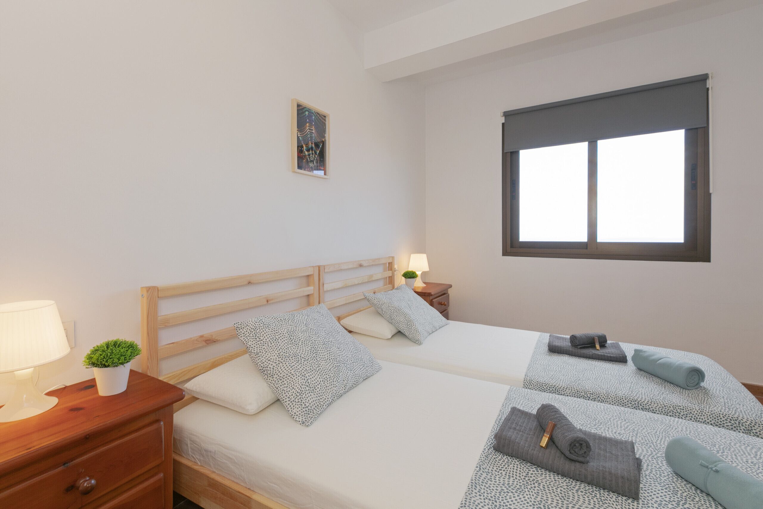 Una cama doble con cabecera de madera, una mesita de noche con una lámpara y una planta pequeña, y una ventana con persiana.