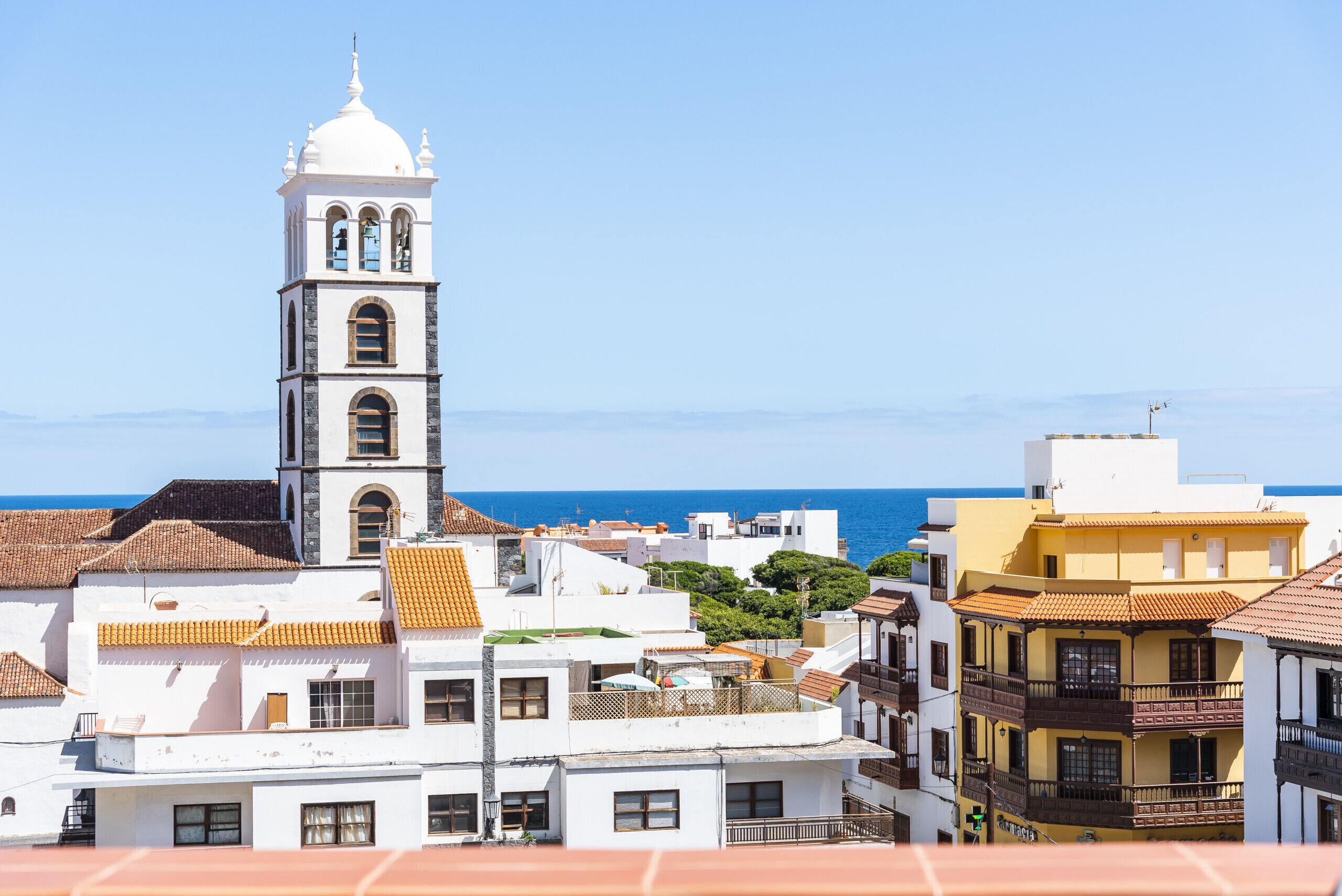 A coastal town with a prominent white bell tower, terracotta-roofed buildings, and a clear view of the sea.