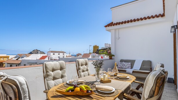 Un área de comedor en la azotea con una mesa preparada para dos personas, rodeada de sillas acolchadas, un frutero y un jarrón con flores.