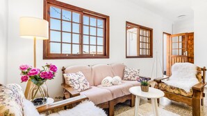 Una sala de estar acogedora con un sofá, un sillón y una mesita. Se observan ventanas grandes con marcos de madera y una lámpara sobre una mesita auxiliar.