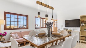 Un comedor con una mesa de madera, sillas blancas y un televisor al fondo.