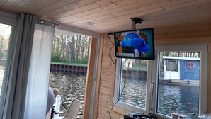 A cabin with a television mounted on the wall, a window overlooking a body of water, and a view of the surrounding forest. - Direkt auf dem Wasser - Un`s Lütt Goldfisch im "haifischbecken" (Röbel/Müritz)