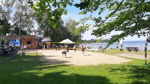 A beach volleyball court with a wooden building in the background, a grassy area, and a body of water. - Direkt auf dem Wasser - Un`s Lütt Goldfisch im "haifischbecken" (Röbel/Müritz)