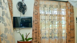 A room with a herringbone-patterned wooden floor, a television mounted on the wall, a wooden chair, a small table with a plant, and patterned curtains. - Bluestone Lounge and Lodge. (Arusha)