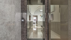 A hallway with marble walls and a glass door leading to a room with a large mirror, a flower arrangement, and a bench. - 3737 La Catedral by Barbarhouse (Mesagne)