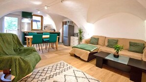 Ein gemütliches Wohnzimmer mit einer Couch, einem Couchtisch und einem grünen Sessel. Es gibt eine Küchenzeile mit einem Esstisch und Stühlen.