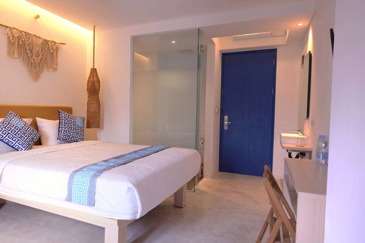 A modern hotel room with a bed, a blue door, and a small bathroom area.