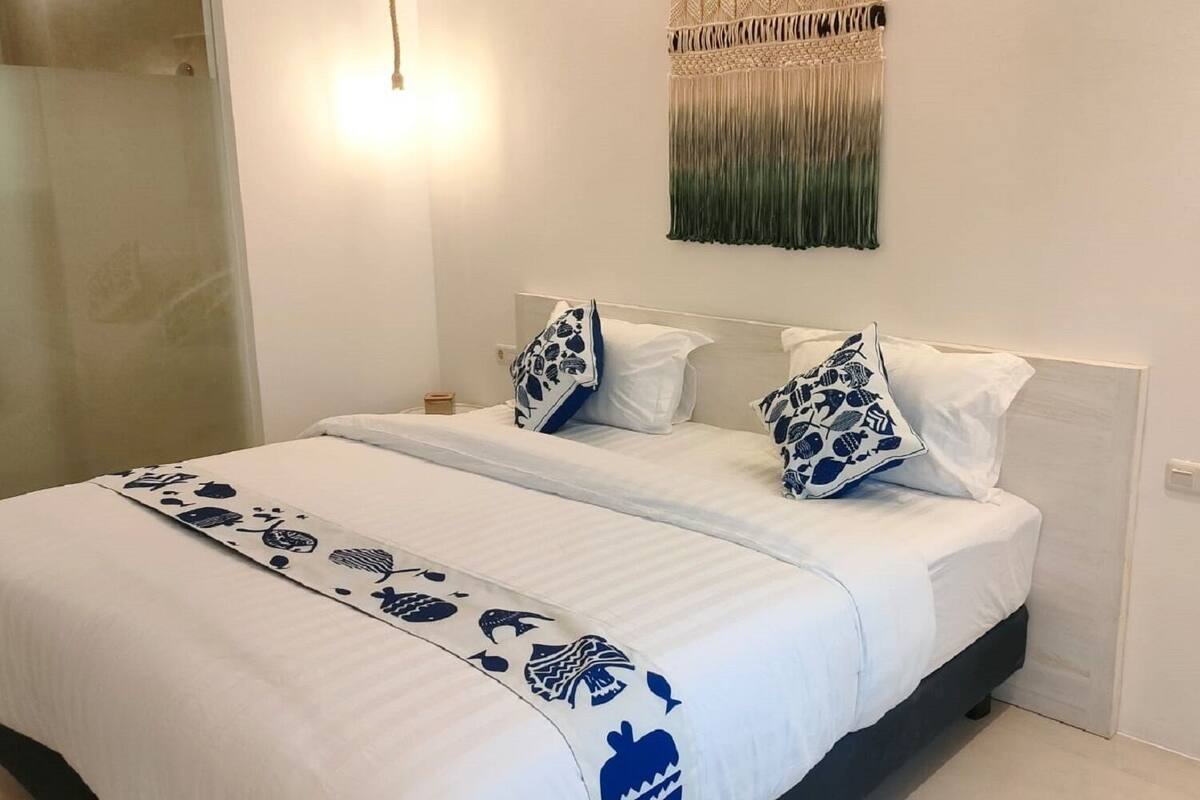 A neatly made bed with a white and blue patterned bedspread, two decorative pillows, and a wall-mounted macrame piece above the bed.