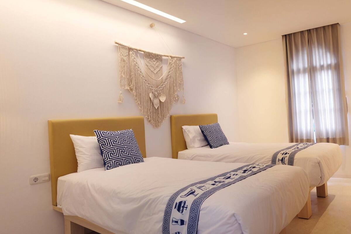 A hotel room with two beds, a wooden headboard, a macrame wall hanging, a large window with curtains, a TV mounted on the wall, and a table with chairs.