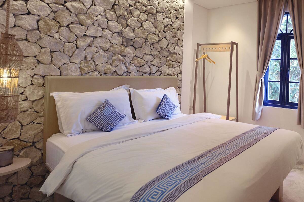A neatly made bed with patterned pillows, a stone wall, a hanging light fixture, a window with curtains, and a small table with a kettle and items.