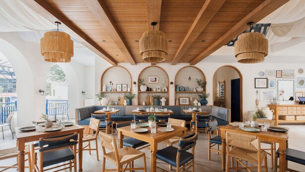 A modern dining area with wooden tables and chairs, hanging wicker chandeliers, and a cozy seating area with a sofa and armchairs. - Little Mykonos (Bandung)