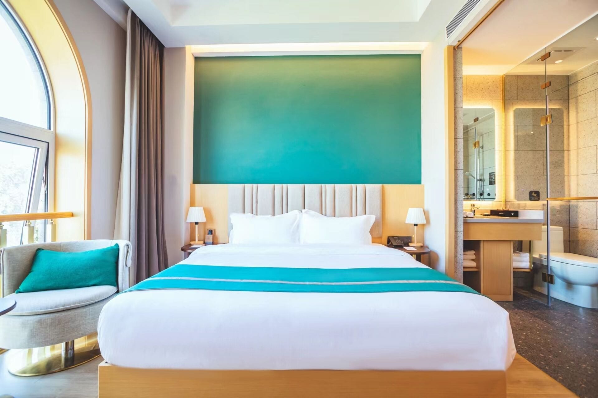 A hotel room with a large bed, a sitting area with a chair, a bathroom with a shower, and a view of the outdoors.