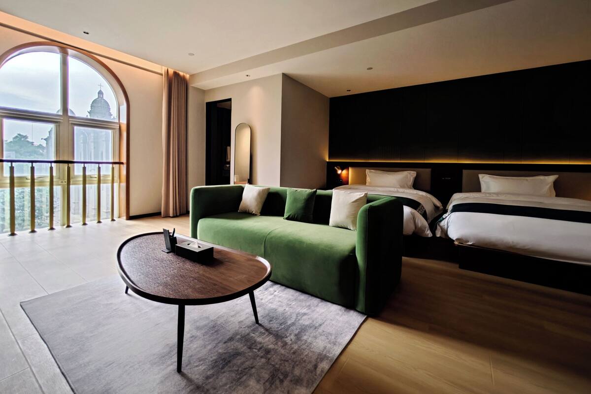 A hotel room with a large window, a green sofa, a bed, and a small table.