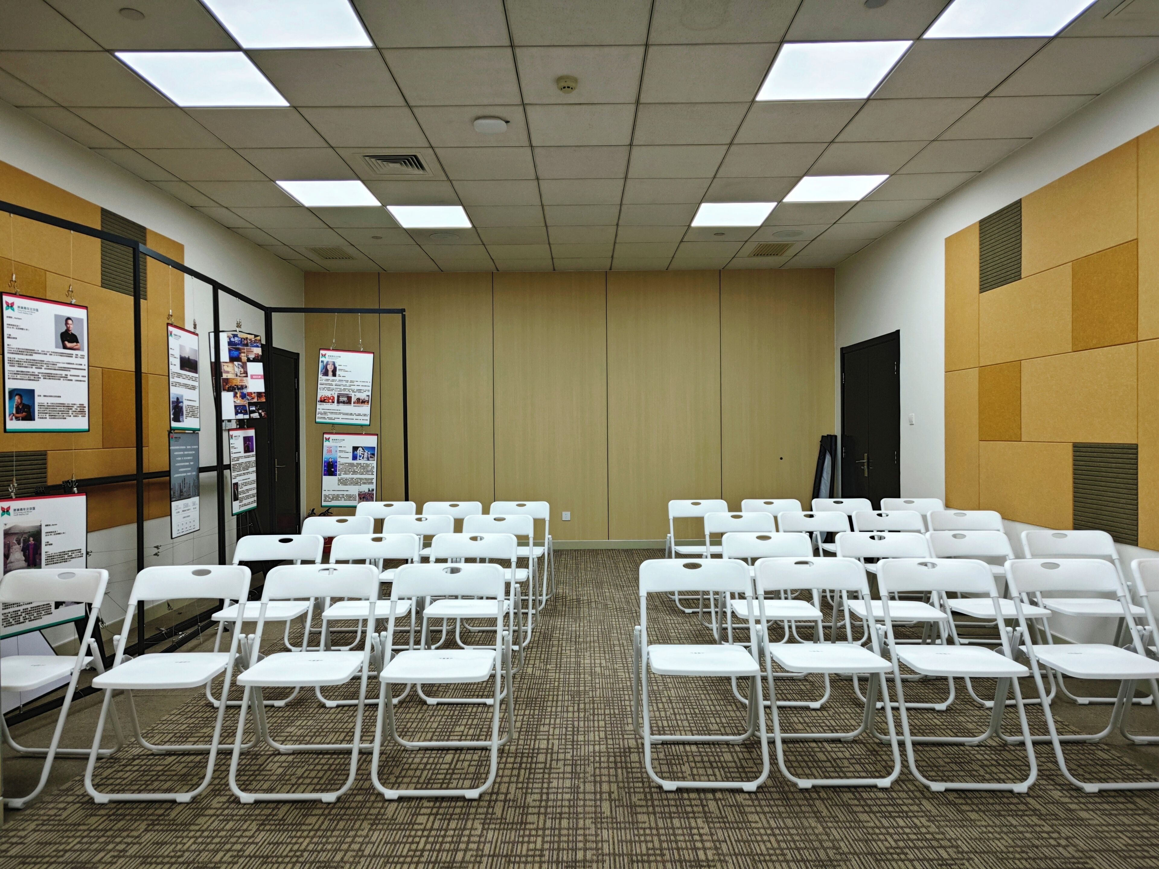 A conference room with white folding chairs arranged in rows, a carpeted floor, and a beige wall with posters.