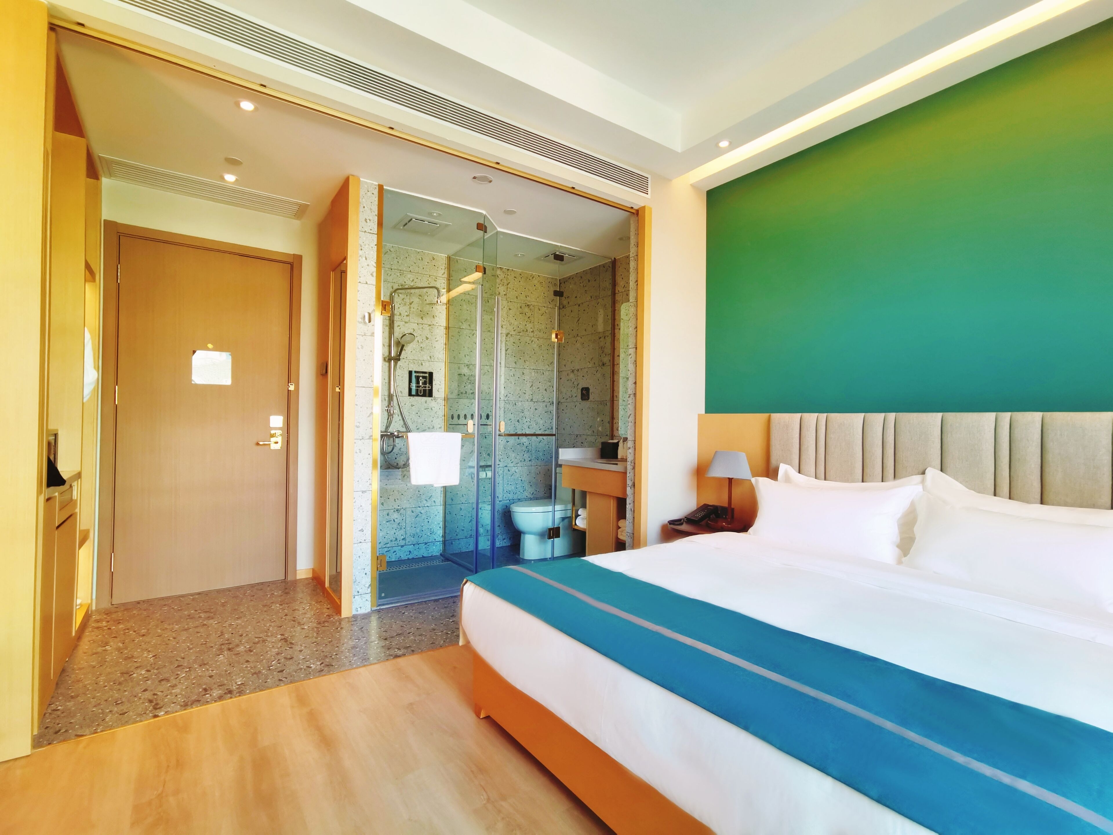 A hotel room with a large bed, a bathroom with a glass shower enclosure, and a green accent wall.