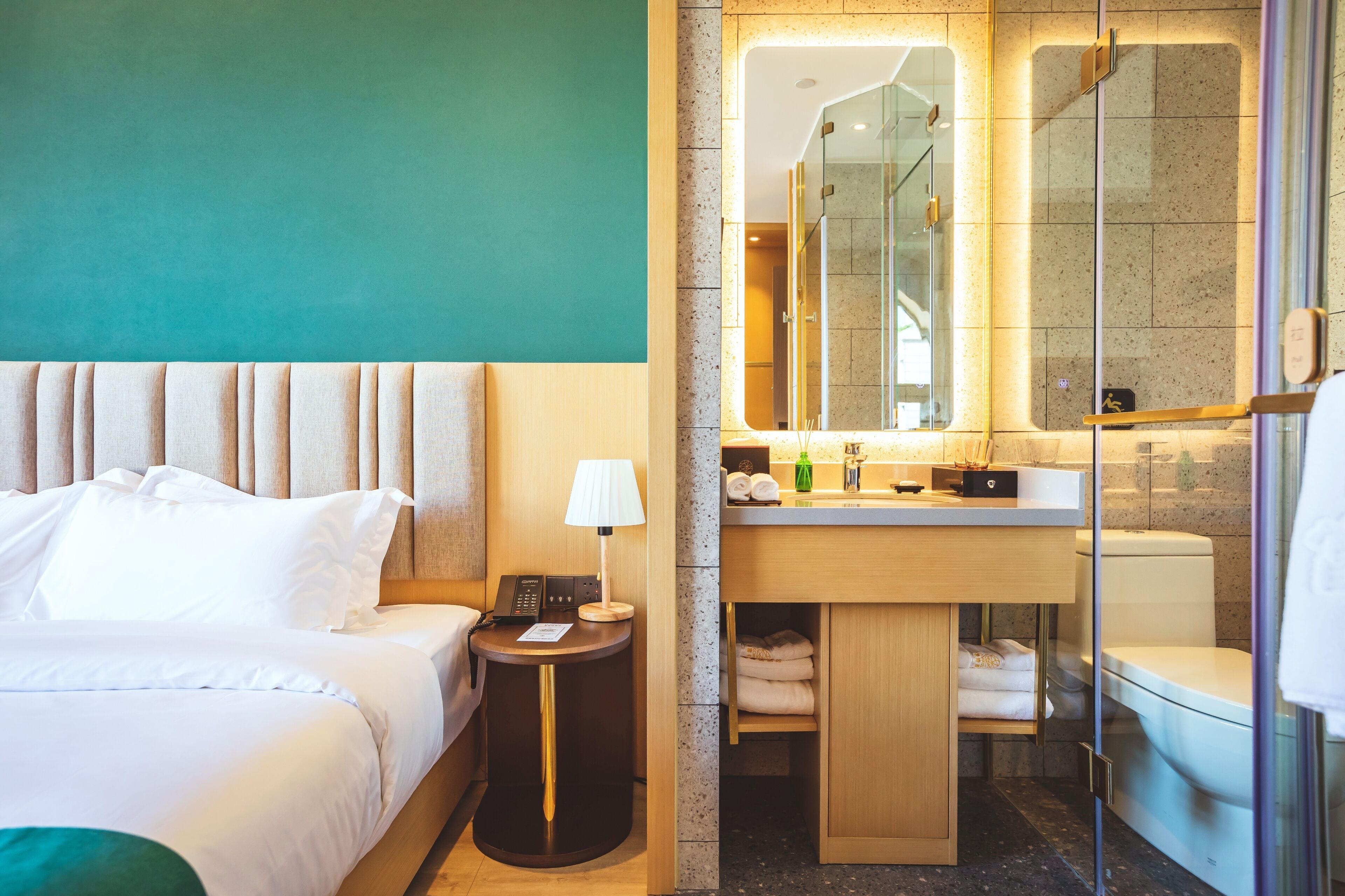 A hotel room with a bed, bedside table, lamp, and a bathroom with a sink, mirror, and toilet.