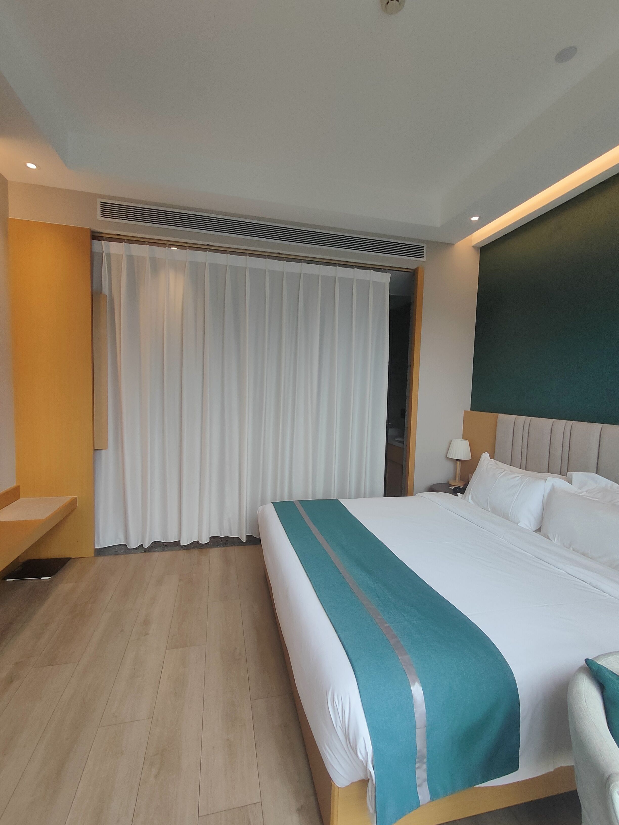 A hotel room with a large bed, a desk, a chair, a nightstand, and a curtain.