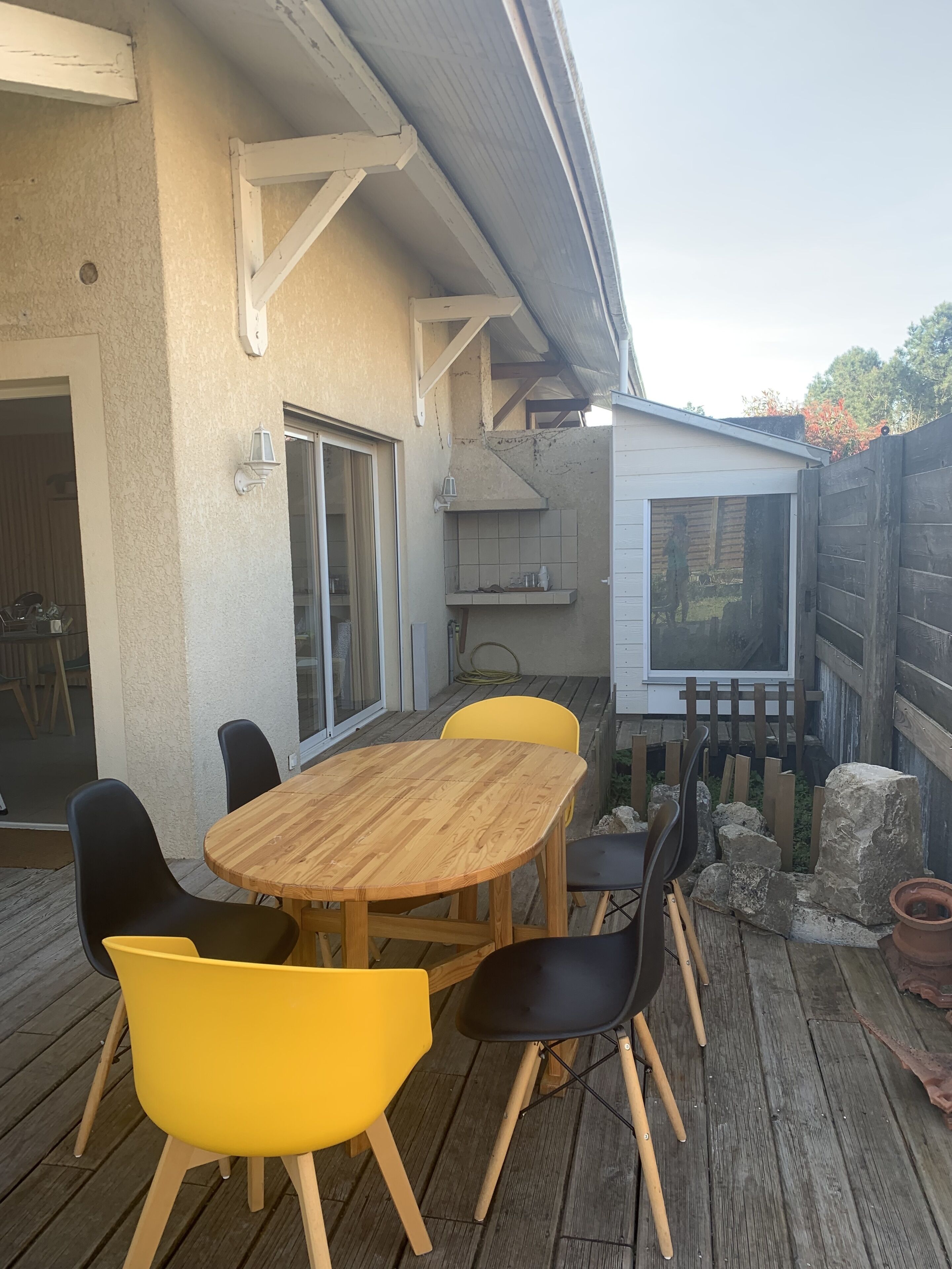 Een terras buiten met een houten tafel en stoelen, een schuifpui en een kleine schuur.