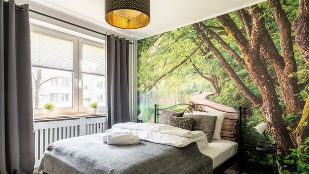 Ein Schlafzimmer mit Bett, einem Fenster mit Vorhängen, einem Heizkörper und einer Wandmalerei von einem Wald.