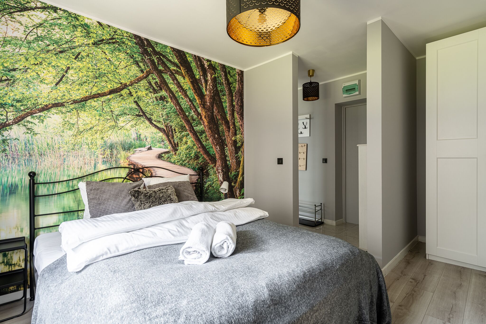A neatly made bed with white linens and a grey blanket, a large forest mural on the wall, a hanging lamp, and a door with a sign.