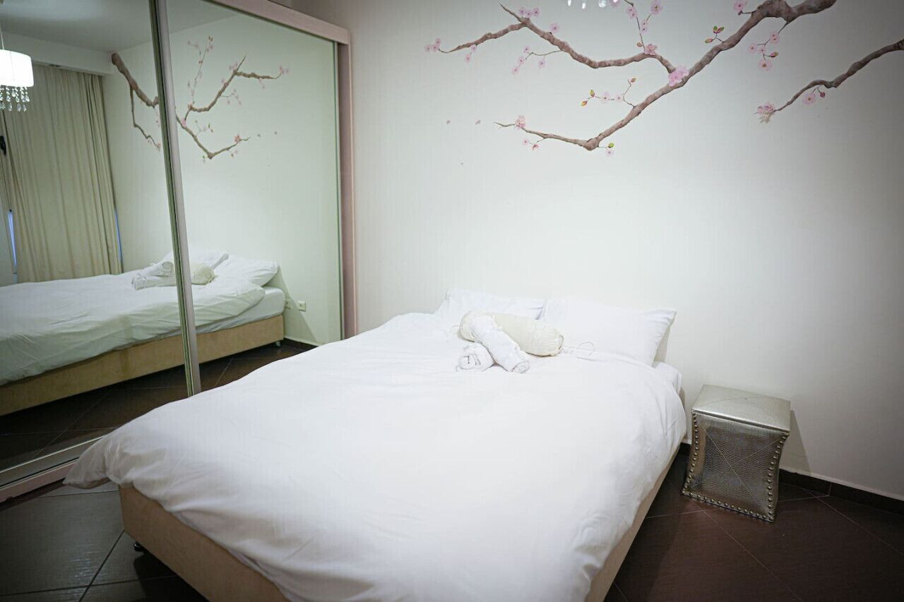 Una camera d'hotel con un letto, un comodino, uno specchio e una decorazione a parete raffigurante un ramo con fiori rosa.