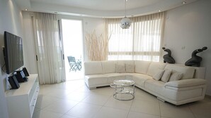 Ein modernes Wohnzimmer mit einem weißen Ecksofa, einem Glastisch und einem Flachbildfernseher auf einer weißen Konsole.