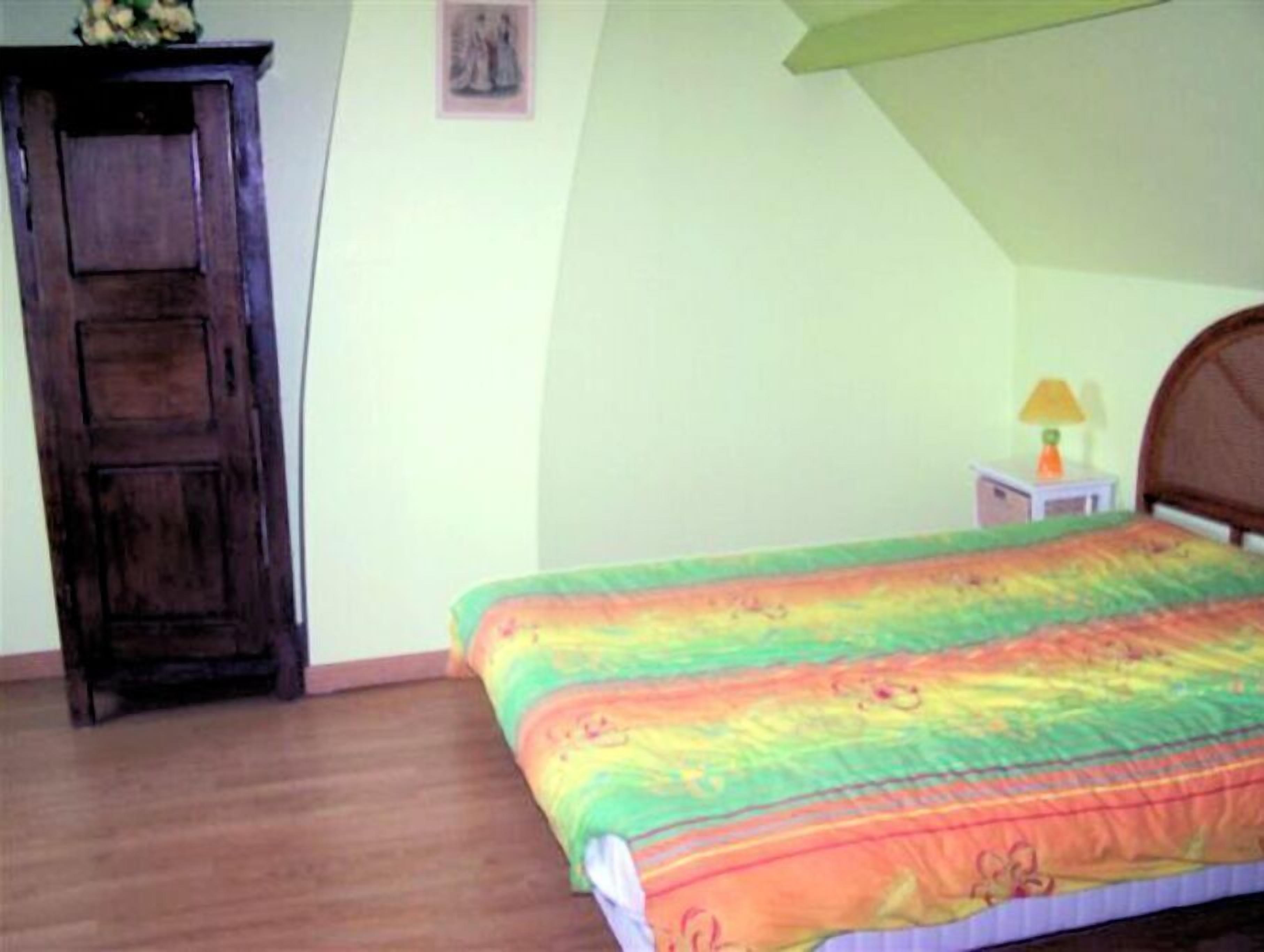 A bedroom with a wooden wardrobe, a bed with a colorful quilt, a nightstand with a lamp, and a framed picture on the wall.
