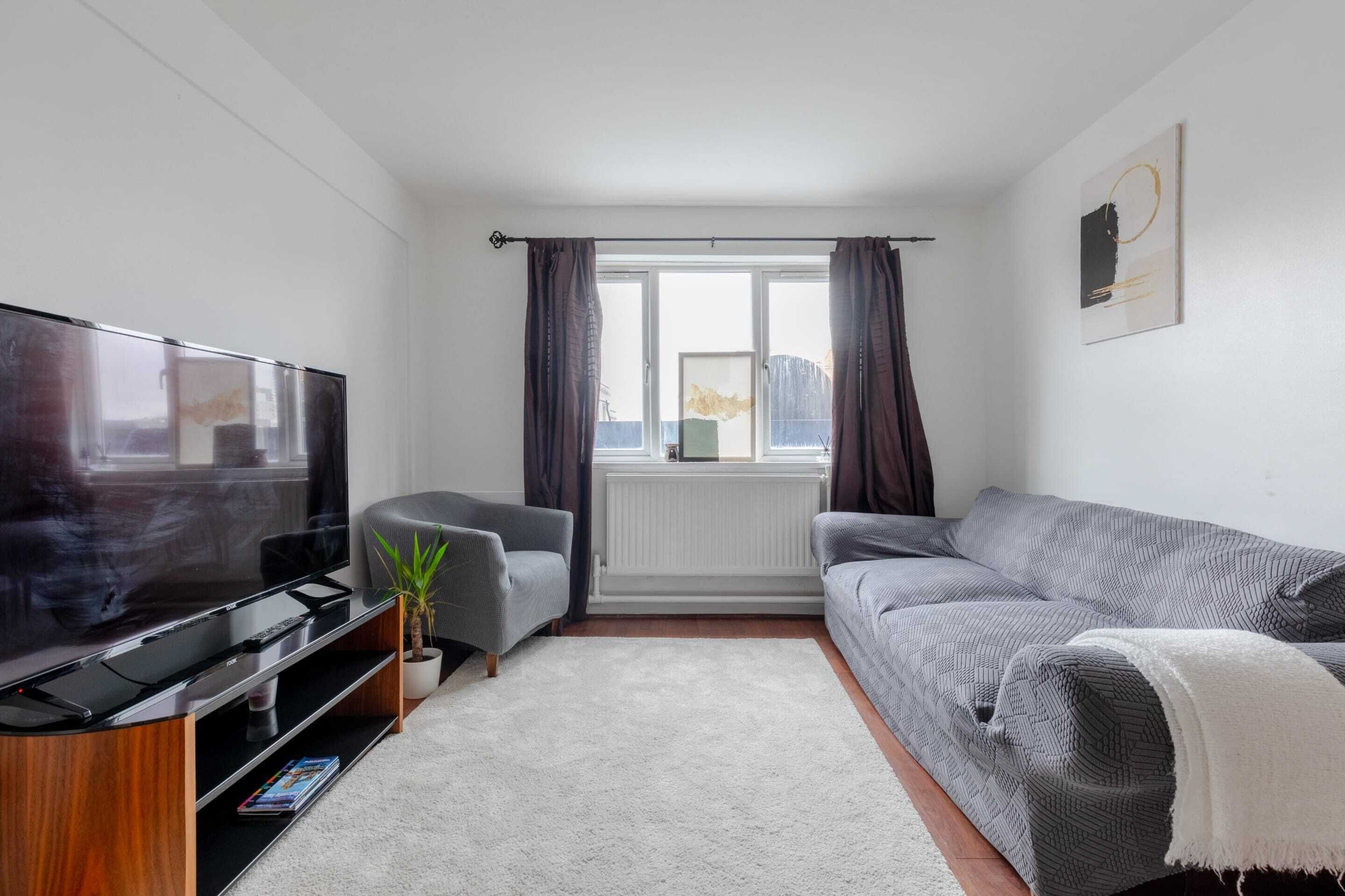 A modern living room with a flat-screen TV, a grey sofa, a grey armchair, a small potted plant, and a window with curtains.